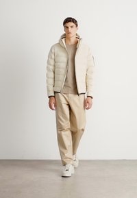 Man loopt vooruit in een beige pufferjack, taupe shirt, beige broek en witte sneakers tegen een witte achtergrond.