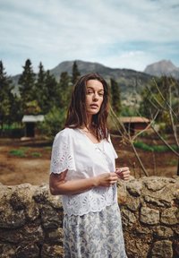 Vit blus med broderade spetsärmar, i kombination med en blommönstrad kjol. Naturlig miljö med en stenmur och grönska i bakgrunden.
