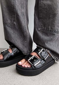 Černé lakované platformové sandály s dvěma přezkami, odpočívající na šedém betonu, spárované s volnými šedými džíny.