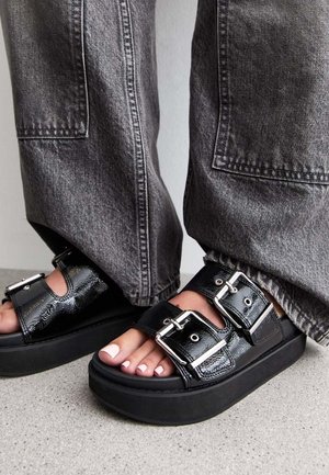 Musta kiilakorkolevy sandaalit, joissa on kaksi solkea, lepäävät harmaalla betonilla, yhdistettyinä tilaviin harmaisiin farkkuroihin.