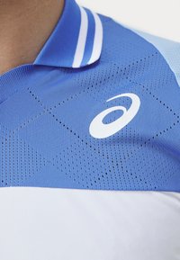 Niebieska i biała koszulka polo sportowa z teksturowanym, przewiewnym górnym fragmentem i jednolitą dolną częścią. Posiada kołnierzyk z dwoma białymi paskami oraz logo.