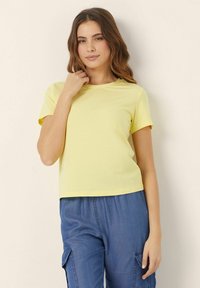 T-shirt giallo a maniche corte con scollatura rotonda; realizzato in un tessuto liscio e leggero. Abbinato a pantaloni cargo blu.