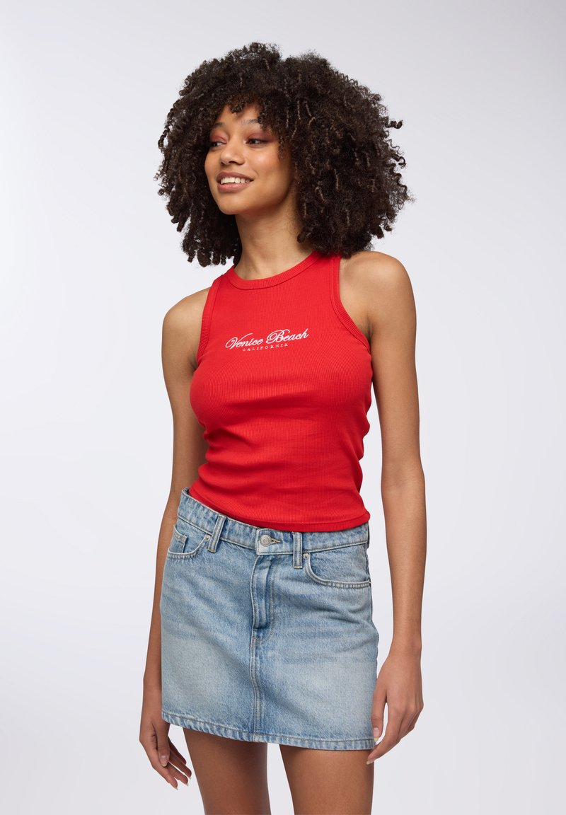 Top a fascia rosso con righe e la frase "Venice Beach California" in bianco, abbinato a una mini gonna in denim azzurro chiaro. Texture liscia, design aderente.