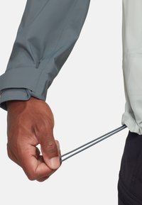 Hand die de zoom van een grijs jack met elastisch aantrekkoordje en trekstopper aanpast tegen een effen achtergrond.