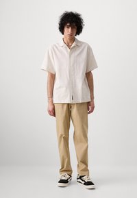 Chemise à manches courtes, beige clair, avec un col, assortie à un pantalon beige et des baskets noir et blanc. Le modèle se tient devant un fond uni.