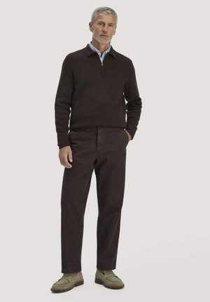 Man staat en draagt een donkerbruine trui met ritssluiting, donkerbruine broek, lichtblauwe overhemdkraag zichtbaar, en beige loafers tegen een effen achtergrond.