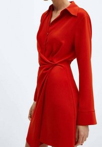 Vestido rojo de camisa, confeccionado en una tela suave, con mangas largas, cuello camisero, cierre frontal de botones y un detalle retorcido en la cintura.