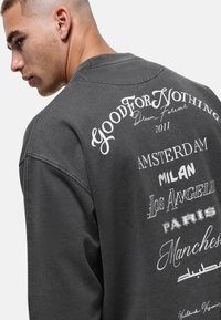 Overmaat grijze sweatshirt met een ronde halslijn, met witte bedrukte tekst van stedenamen en decoratieve elementen op de achterkant.