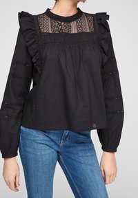 Schwarze Bluse mit Rüschenausschnitt und -ärmeln, verziert mit Spitzenpaneelen und Stickereien. Kombiniert mit blauen Jeans.