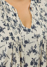 Nahaufnahme einer cremefarbenen Bluse mit navyblauem Blumenmuster, gefältelten Details und kleinen Pailletten im Bereich des Ausschnitts und der Brust.