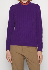 Lila kabelstickad tröja med ribbad halsringning och fåll, kombinerad med bruna skräddarsydda byxor. Tröjan har ett strukturerat mönster.