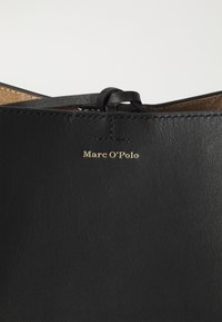 Prodotto in pelle nera con superficie testurizzata. Presenta un piccolo nodo in pelle e il logo dorato "Marc O'Polo" impresso sul davanti.