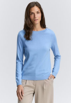 Pull bleu clair en tissu doux, avec un col rond et des manches longues. Ourlets et poignets côtelés, porté avec un pantalon beige.