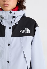 Asmuo vilkintis šviesiai pilką ir juodą „North Face“ striukę su kapišonu ir matomu prekės ženklu ant krūtinės, po ja matosi raudona merino vilnos apykaklė.