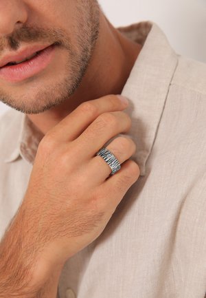 Anello in argento con texture, indossato su una mano, caratterizzato da un design ondulato; sullo sfondo si intravede una camicia in lino di colore chiaro.
