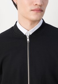 Μαύρο μπόμπορτζ jacket με λεία υφή, διαθέτει μπροστινό φερμουάρ και άσπρη πουκάμισο από κάτω. Κοντινή λήψη του λαιμού.