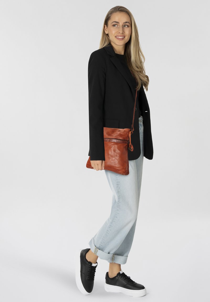 Giovane donna che indossa un blazer nero, jeans arrotolati blu chiaro, sneakers nere e porta una borsa a tracolla in pelle marrone, sorridendo di lato.