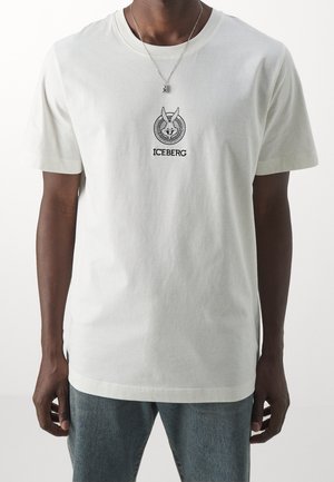 Mand iført en hvid T-shirt med sort tegneserie-kaninlogo og ordet "ICEBERG" under, parret med blå jeans.