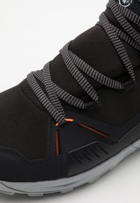 Stivale sportivo nero con lacci grigi, dotato di una tomaia strutturata, dettagli arancioni e una suola in gomma robusta con motivo a rilievo.