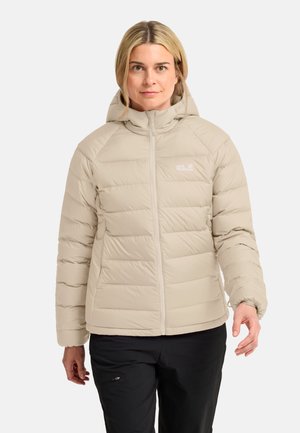 Vrouw draagt een beige gewatteerde jas met capuchon en zwarte broek, staand tegen een effen lichte achtergrond.