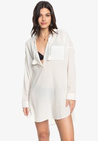 Camicia leggera, bianca, a maniche lunghe con un taglio rilassato, scollatura a V profonda, taschino sul petto e dettagli a frange sul colletto.