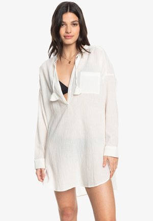 Camicia leggera, bianca, a maniche lunghe con un taglio rilassato, scollatura a V profonda, taschino sul petto e dettagli a frange sul colletto.