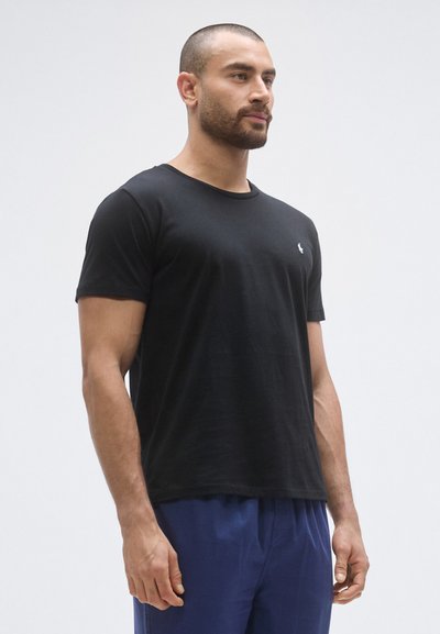 Črna bombažna majica s kratkimi rokavi, okroglim izrezom in majhnim logotipom na prsih. Prikazana je oprijeta silhueta s modrimi hlačami.