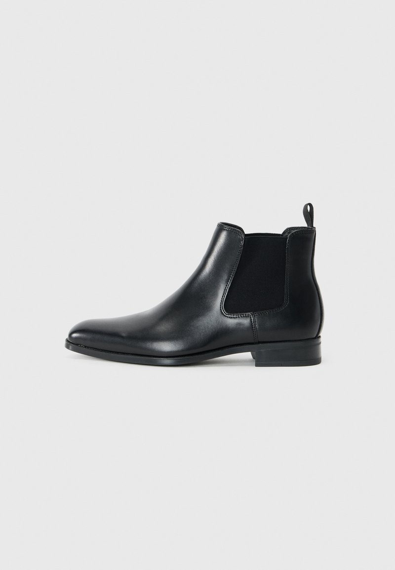 Bota Chelsea de cuero negro con paneles elásticos en los laterales, puntera redondeada, pequeño tirador en la parte trasera y tacón bajo apilado. Textura suave.