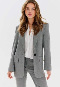 Sivý a čierny károvaný oblek s priliehavým sako a zodpovedajúcimi nohavicami. Tkanina má textúrovaný vzor, obsahuje zárezy na klopách a jeden gombík.