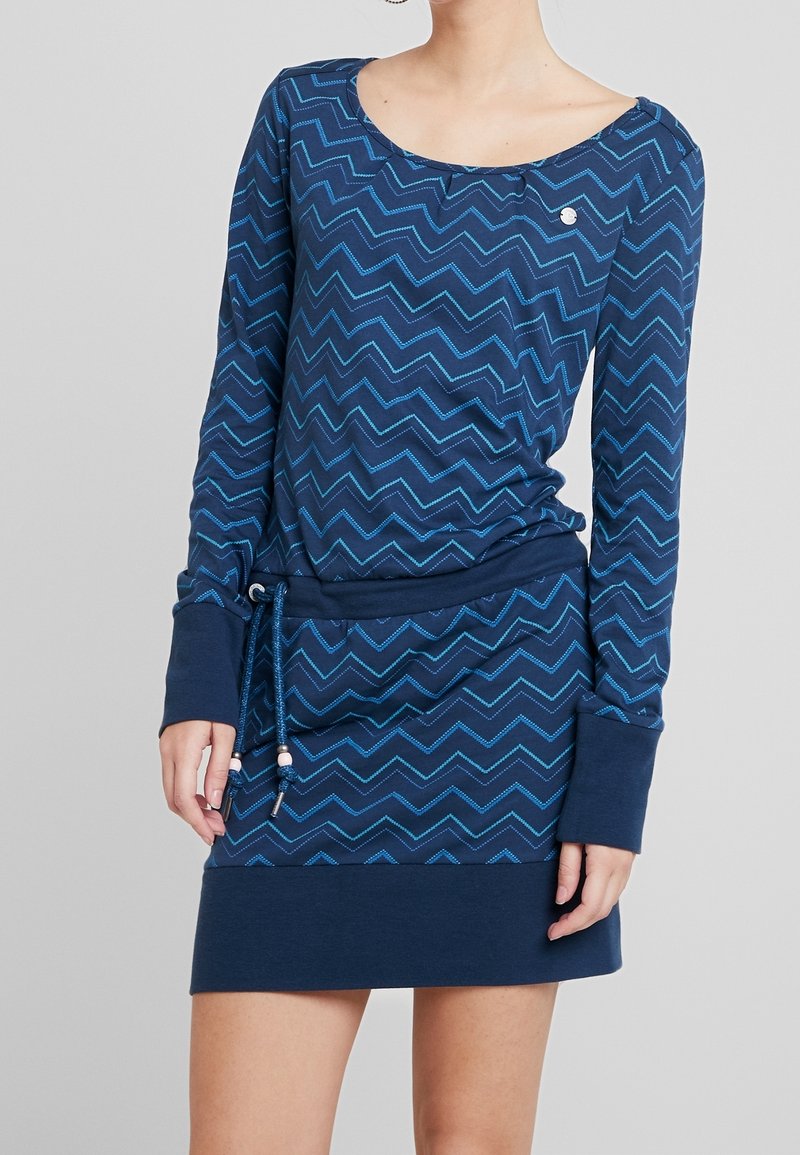 Femme portant une robe bleue à manches longues avec un motif zigzag bleu clair et une taille à cordon, debout devant un fond uni gris clair.