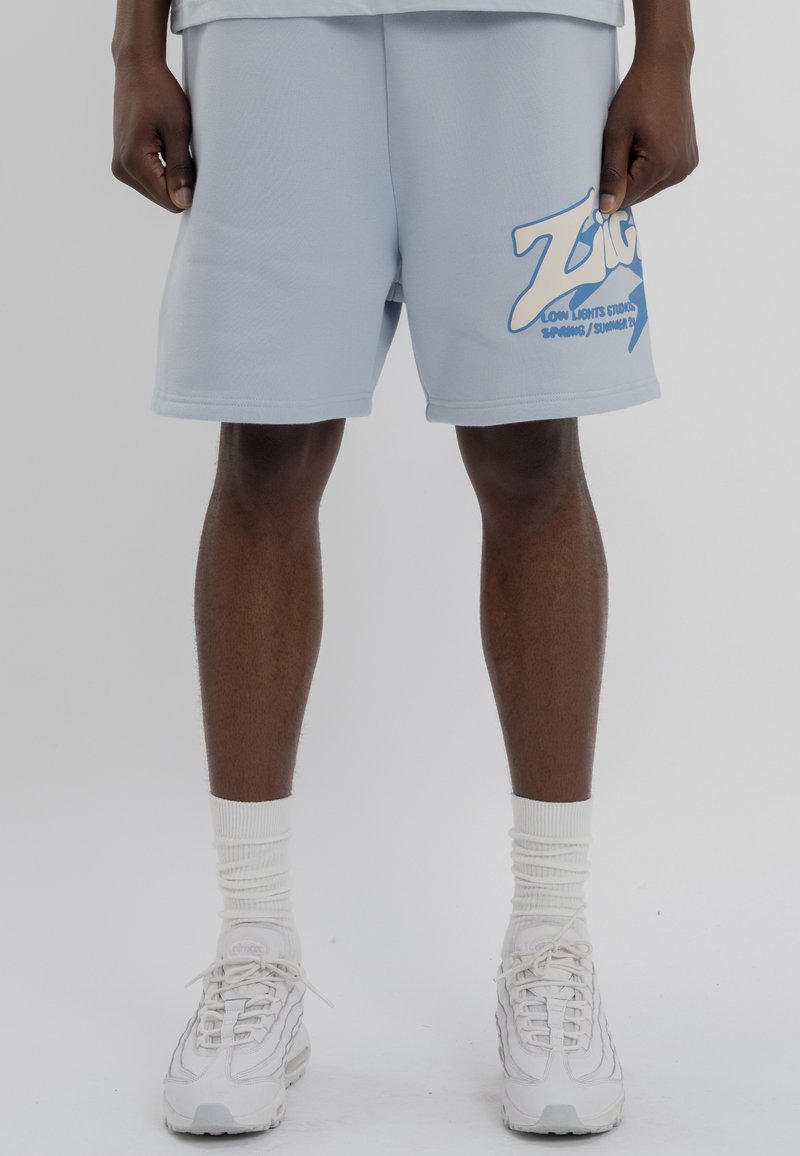 Světle modré kraťasy s hladkou texturou, na levém stehně s velkým bílým grafickým logem. Doladěno s bílými teniskami a ponožkami.