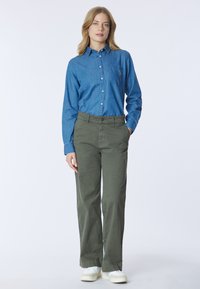 Donna in piedi con le mani nelle tasche, indossa una camicia blu con bottoni, pantaloni verde oliva e sneakers bianche su sfondo bianco.