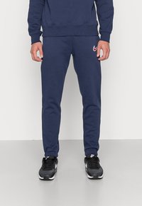 Man klädd i marinblå träningsoverallsbyxor och matchande sweatshirt med röd Nike-logga, står inomhus på ett ljusgrått golv.