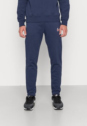 Man in eenmarineblauwe trainingsbroek en bijpassende sweatshirt met rood Nike-logo, staande binnen op een lichtgrijze vloer.