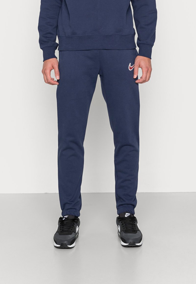 Man klädd i marinblå träningsoverallsbyxor och matchande sweatshirt med röd Nike-logga, står inomhus på ett ljusgrått golv.