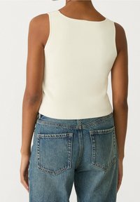 Person som bär en ärmlös vit topp och blå denimjeans, visad från ryggen mot en ljus enfärgad bakgrund.