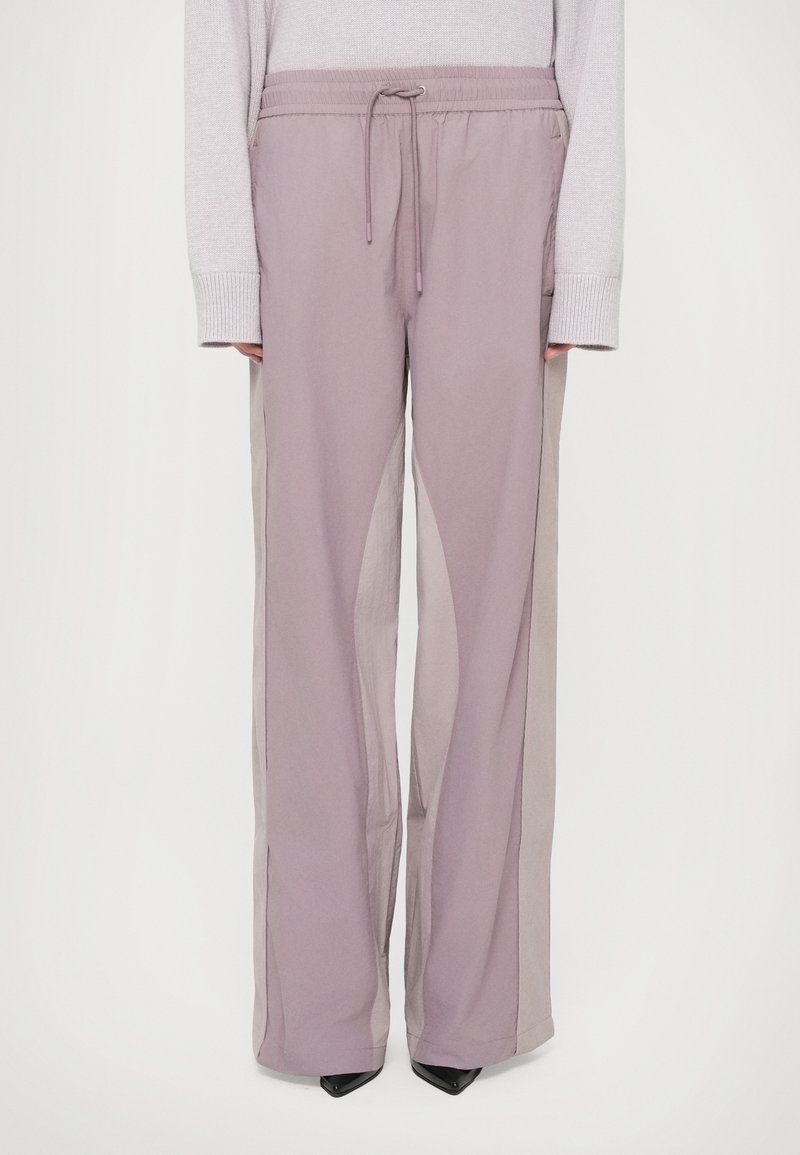 Calças largas de cor lilás claro com cintura elástica. Apresenta painéis laterais transparentes e uma textura suave. Combinadas com sapatos de salto pretos.