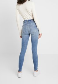 Jeans ajustados de un azul claro, hechos de mezclilla, con una cintura alta, bolsillos traseros y una textura suave. Llevados con una camiseta de manga larga y zapatillas blancas.