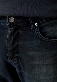 Jeans de denim azul oscuro con un ajuste slim, que cuentan con un botón de metal plateado y remaches, detalles de costura visibles y un bolsillo frontal.