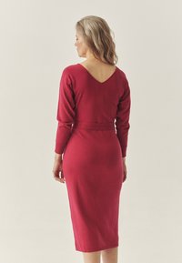 Rød strikket kjole med lange ærmer, V-udskæring bagpå, tætsiddende talje og mellemlangt fald. Glat tekstur, minimal dekoration, elegant silhuet.