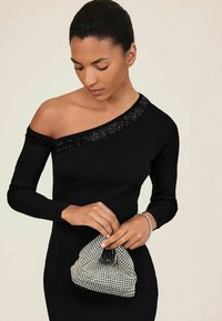 Robe noire à côtes avec une épaule ornée de sequins, accompagnée d'une pochette argentée ornée de bijoux. Bracelet couleur chair au poignet.