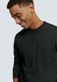 Pull en tricot gris foncé avec une finition texturée, doté d'un col côtelé et d'un petit logo hexagonal au centre.