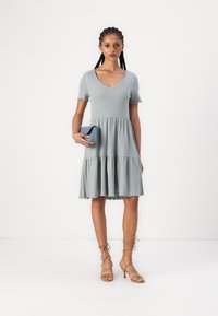 Vestido azul-claro com detalhes em nervuras, decote em V, mangas curtas, saia em camadas e barra em forma de concha; combinado com uma clutch azul e sapatos de salto nude com tiras.