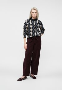 Moteris stovi su argyle raštu sijonu, juoda apykakle marškinėliais, tamsiomis plačiomis kelnėmis ir bordo spalvos baleto bateliais prieš paprastą foną.