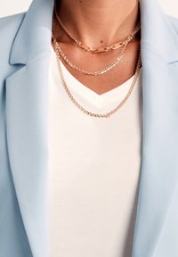 Ljusblå kavaj med slät textur, lager på en vit V-ringad skjorta. Tre halsband i guldton med varierande länkar pryder halsen.