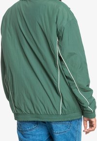 Zielona lekka kurtka o teksturowanym wykończeniu, z białym przeszyciem wzdłuż rękawów i marszczonym dołem, noszona na niebieskich dżinsach.