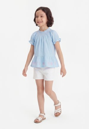 Χαμογελαστό παιδί που περπατά, φοράει γαλάζιο μπλουζάκι με κοντά μανίκια και βολάν, λευκό σορτς και λευκά σανδάλια σε απλό λευκό φόντο.