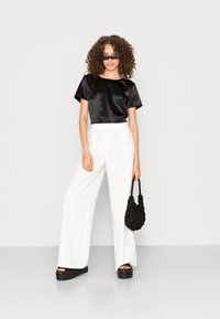 Top preto de cetim com mangas curtas, calças brancas de perna larga, sandálias de plataforma pretas e uma mala texturizada preta. Modelo com cabelo encaracolado e óculos de sol.
