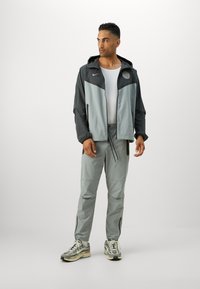 Gray Nike jacket with a black accent, hood, front zipper, and logo. Paired with gray pants and gray sneakers featuring a pattern.