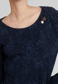 Mörkblå topp med rundad halsringning som har ett fläckigt mönster i ljusblått. Inkluderar dekorativa knappar i trä och silver.